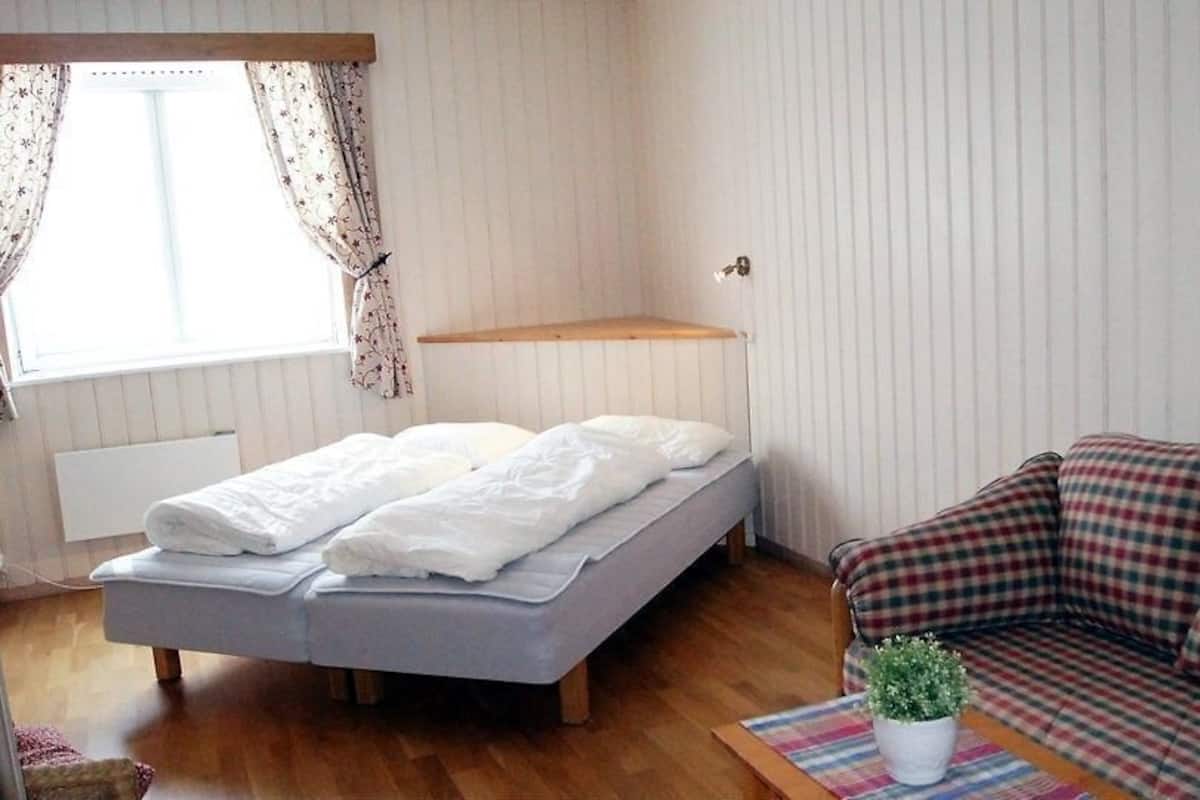 Ett sovrum med en säng, ett sängbord, ett fönster med gardiner, ett trägolv, en rutig soffa och en tavla på väggen.