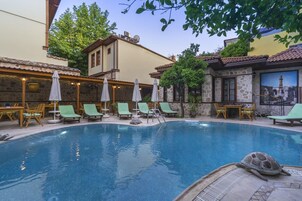 Une piscine entourée de chaises longues et de parasols, le tout bordé de murs en pierre et surmonté d’un toit en tuiles.