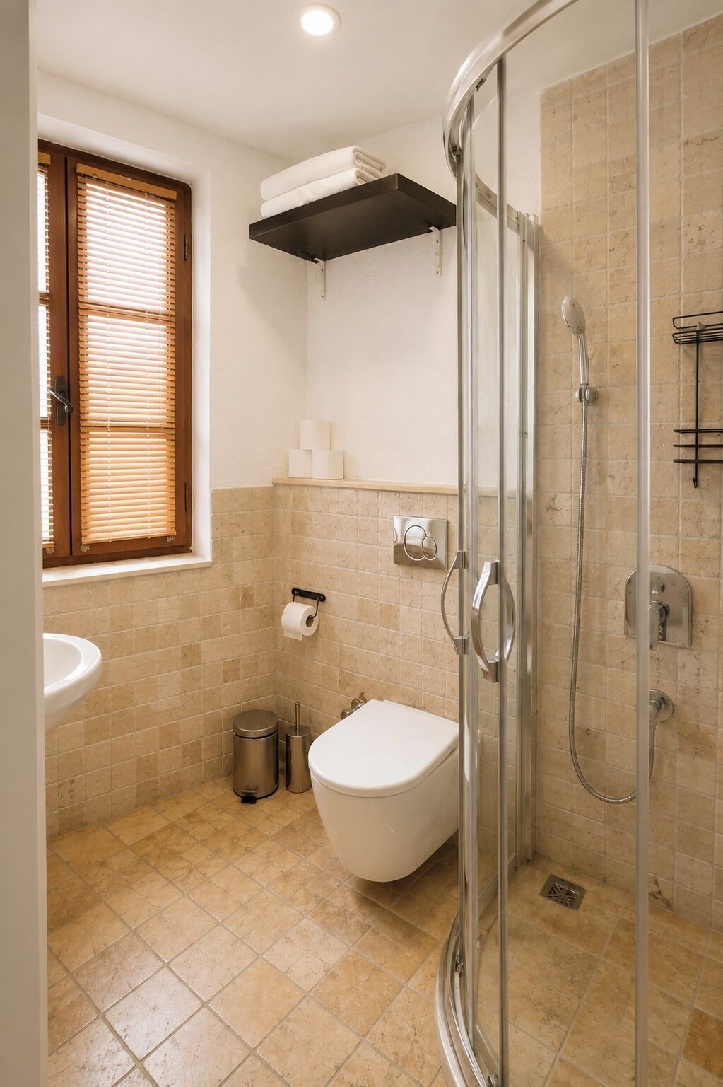 Un baño con cabina de ducha de vidrio, inodoro, lavamanos y ventana con persianas.