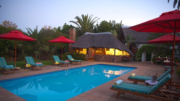 A swimming pool with lounge chairs and red umbrellas, surrounded by greenery and a building with a thatched roof. - Easy Five (Cape Town)