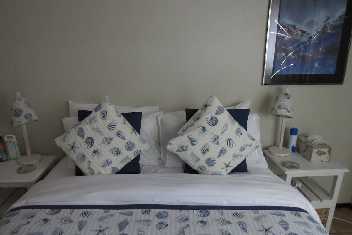 A bed with a blue and white shell pattern bedspread, two matching pillows, a nightstand with a lamp, and a framed picture on the wall.