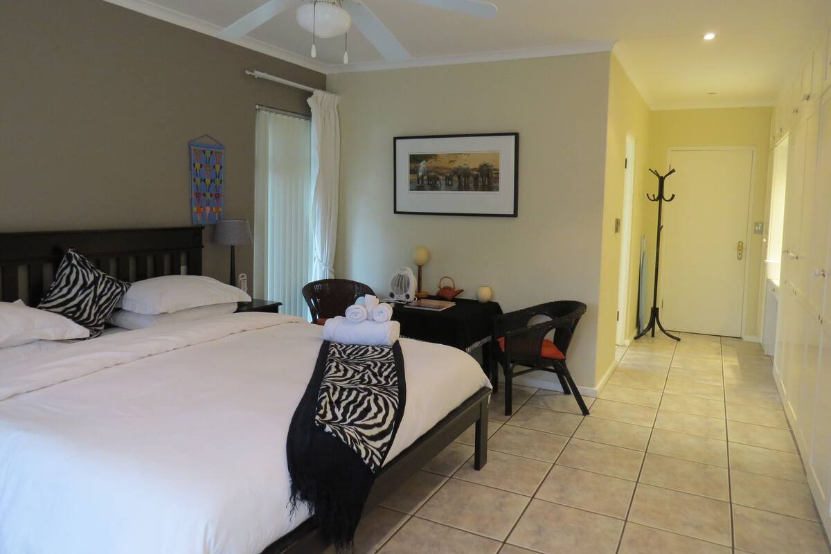 A hotel room with a bed, a desk, a chair, a coat rack, a ceiling fan, and a framed picture on the wall.