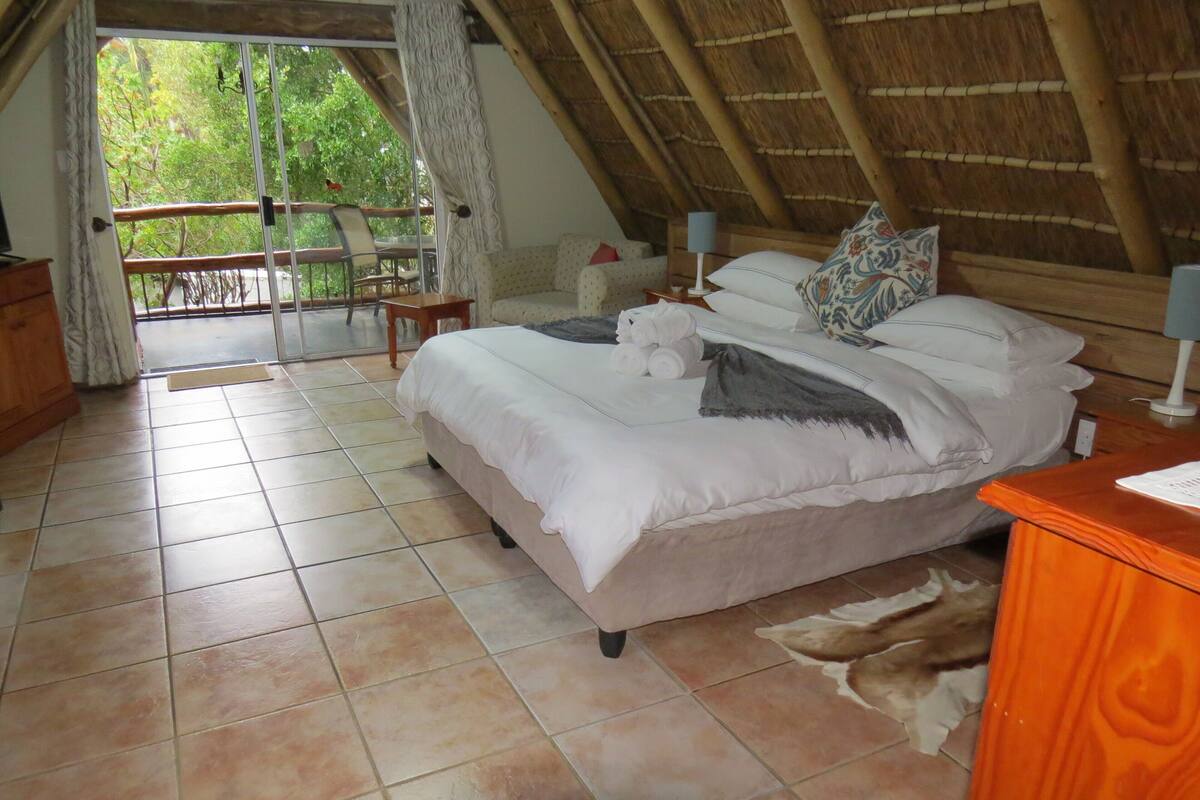 A bedroom with a bed, a wooden nightstand, a chair, and a view of greenery outside.