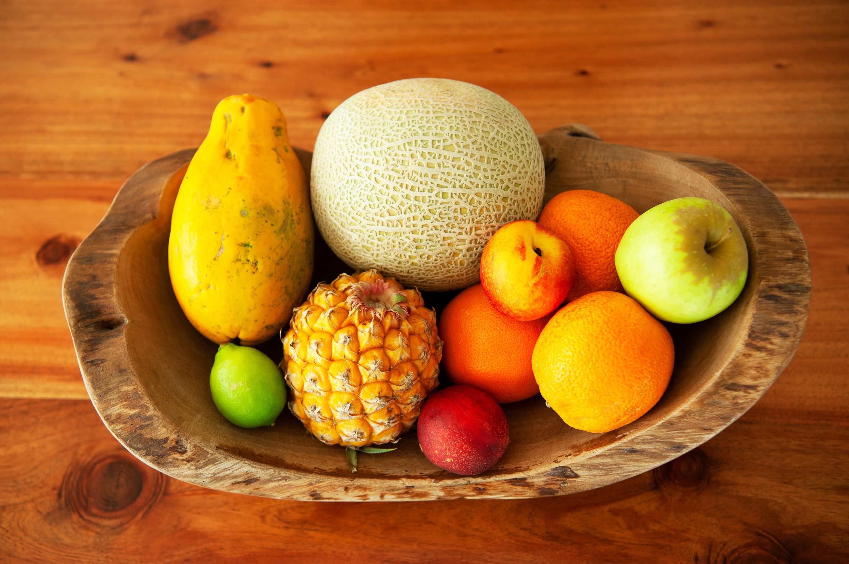 Un cuenco de madera con una variedad de frutas, como melón, piña, naranjas y un melocotón.