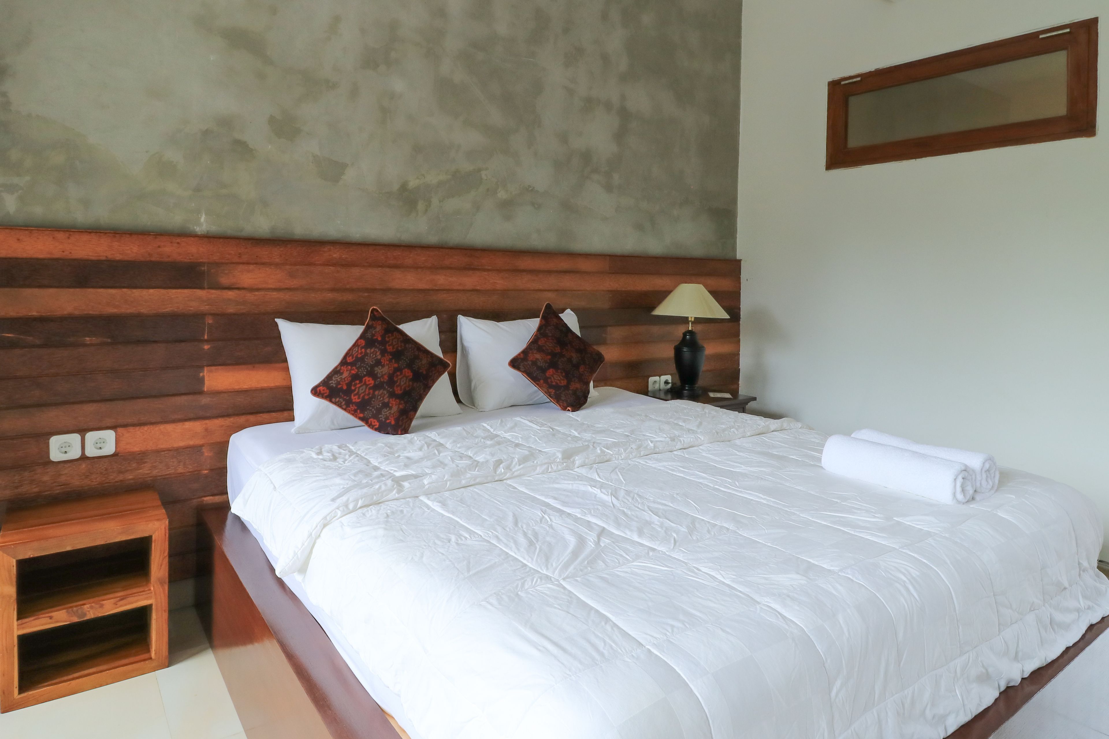 Una cama bien hecha con ropa blanca y almohadas decorativas, una mesita de noche de madera con una lámpara y una pared sencilla con un espejo.