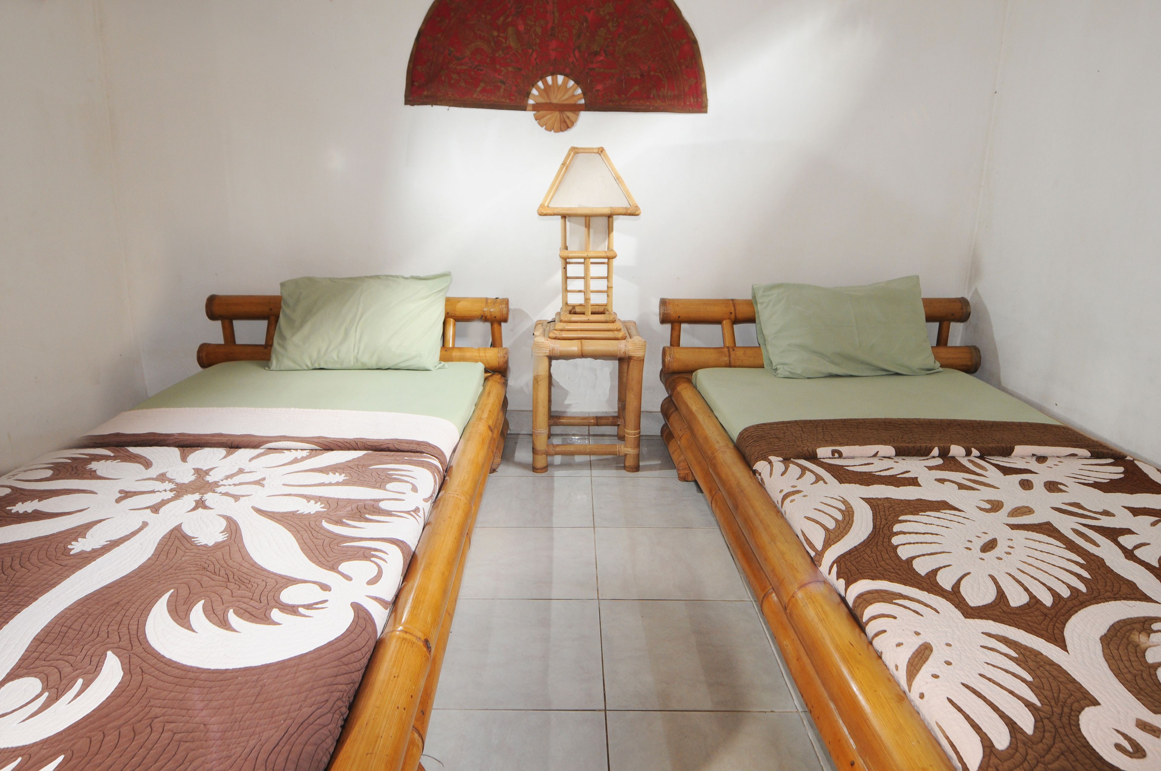 A room with two wooden beds, each with a patterned bedspread, a small wooden nightstand with a lamp, and a red decorative hanging above the nightstand.