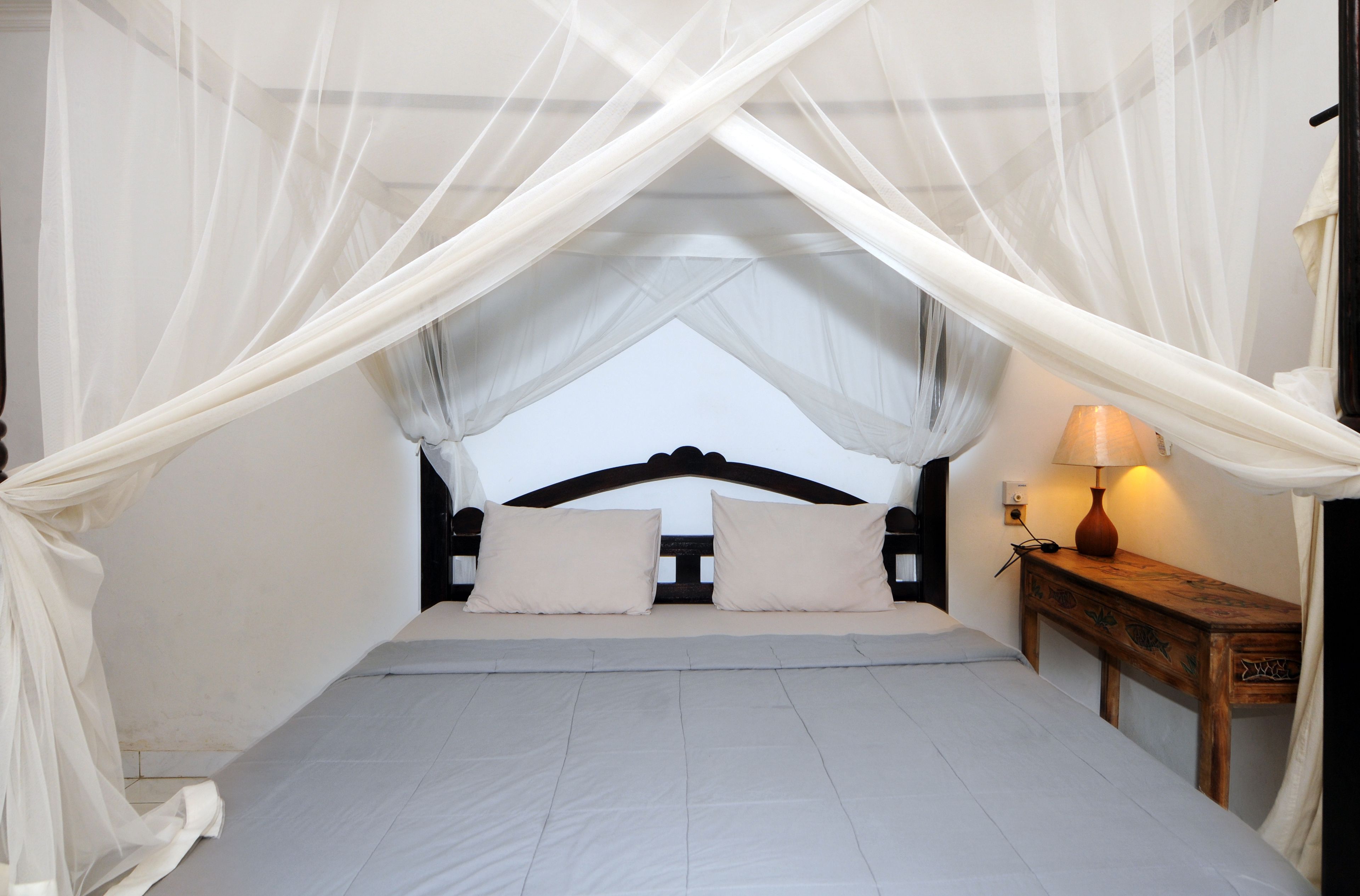 A bedroom with a bed, white curtains, a bedside table with a lamp, and a view of the sky.