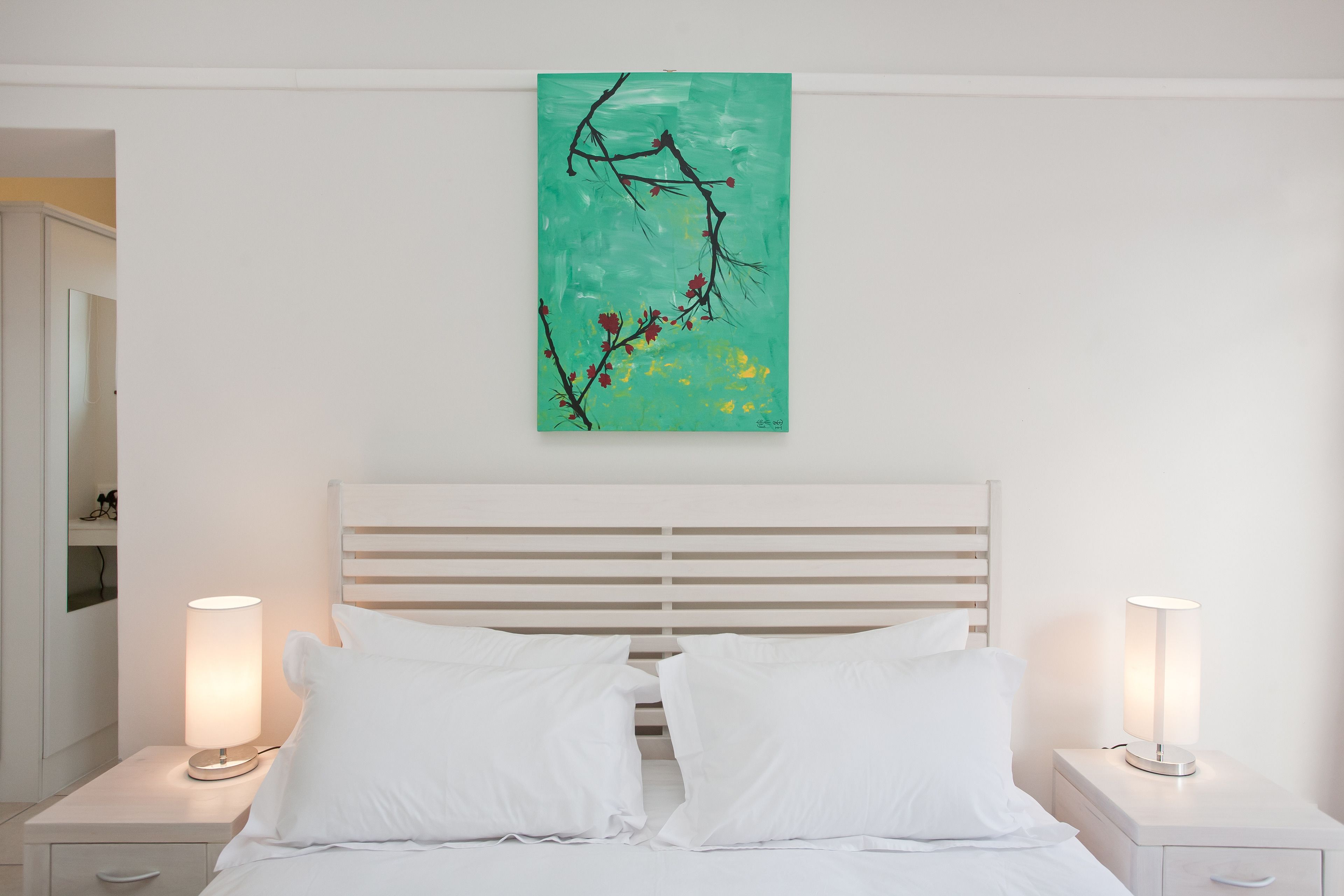 A bedroom with a bed, two bedside tables with lamps, a white headboard, and a wall-mounted painting.