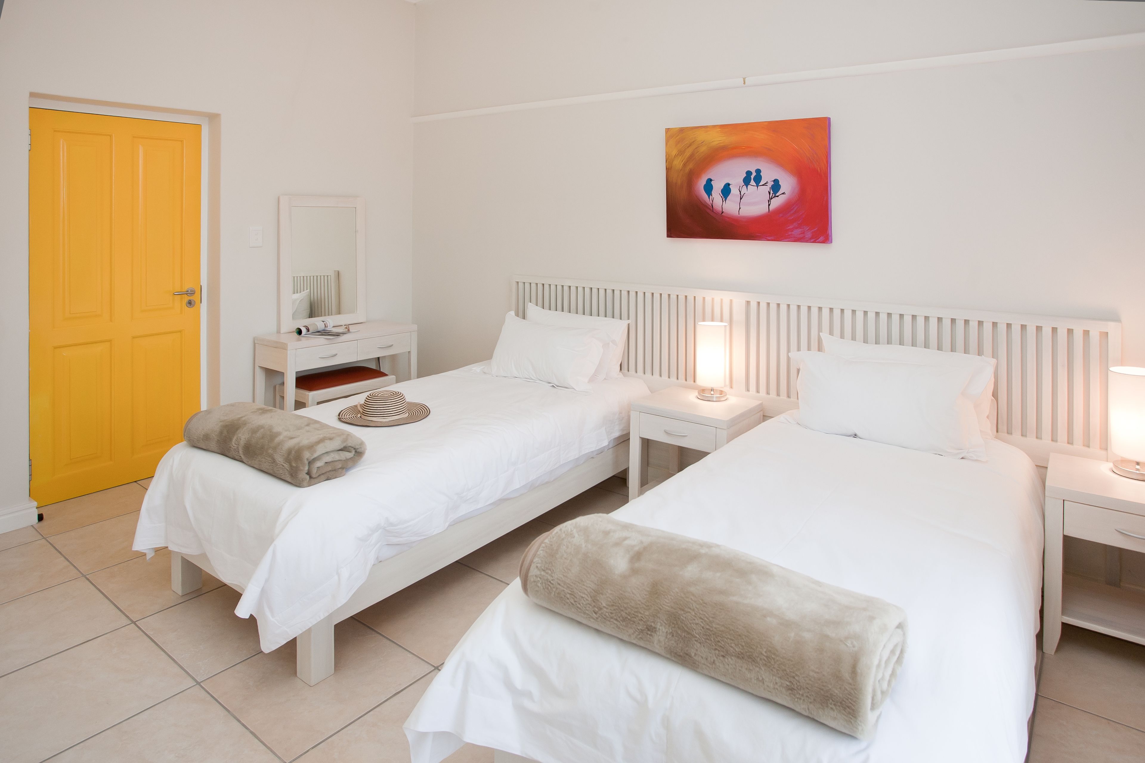 Habitación de hotel con dos camas, una mesita de noche con lámpara, un espejo y un cuadro en la pared.