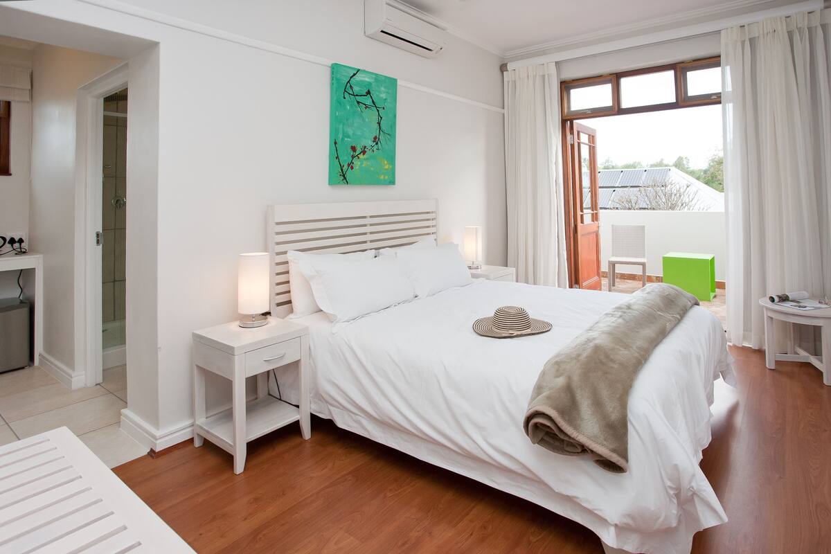 A neatly arranged bedroom with a bed, bedside tables, a nightstand, a window with curtains, and a green wall art piece.