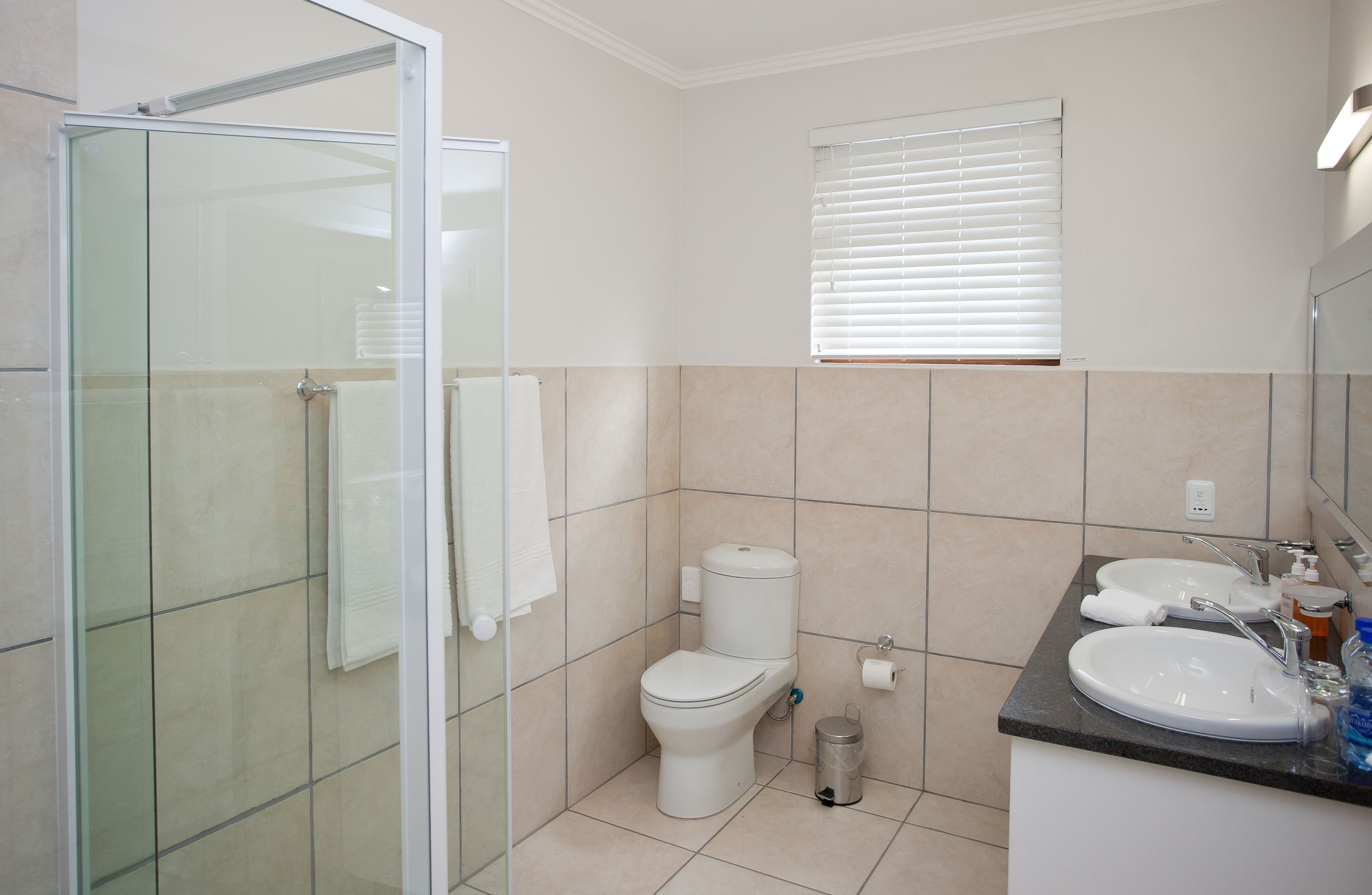 A bathroom with a glass shower enclosure, a toilet, a sink with a mirror, and a window with blinds.