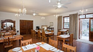 A dining area with wooden tables and chairs, ceiling fans, chandeliers, and large windows with curtains. - The Queen of Calitzdorp (Calitzdorp)