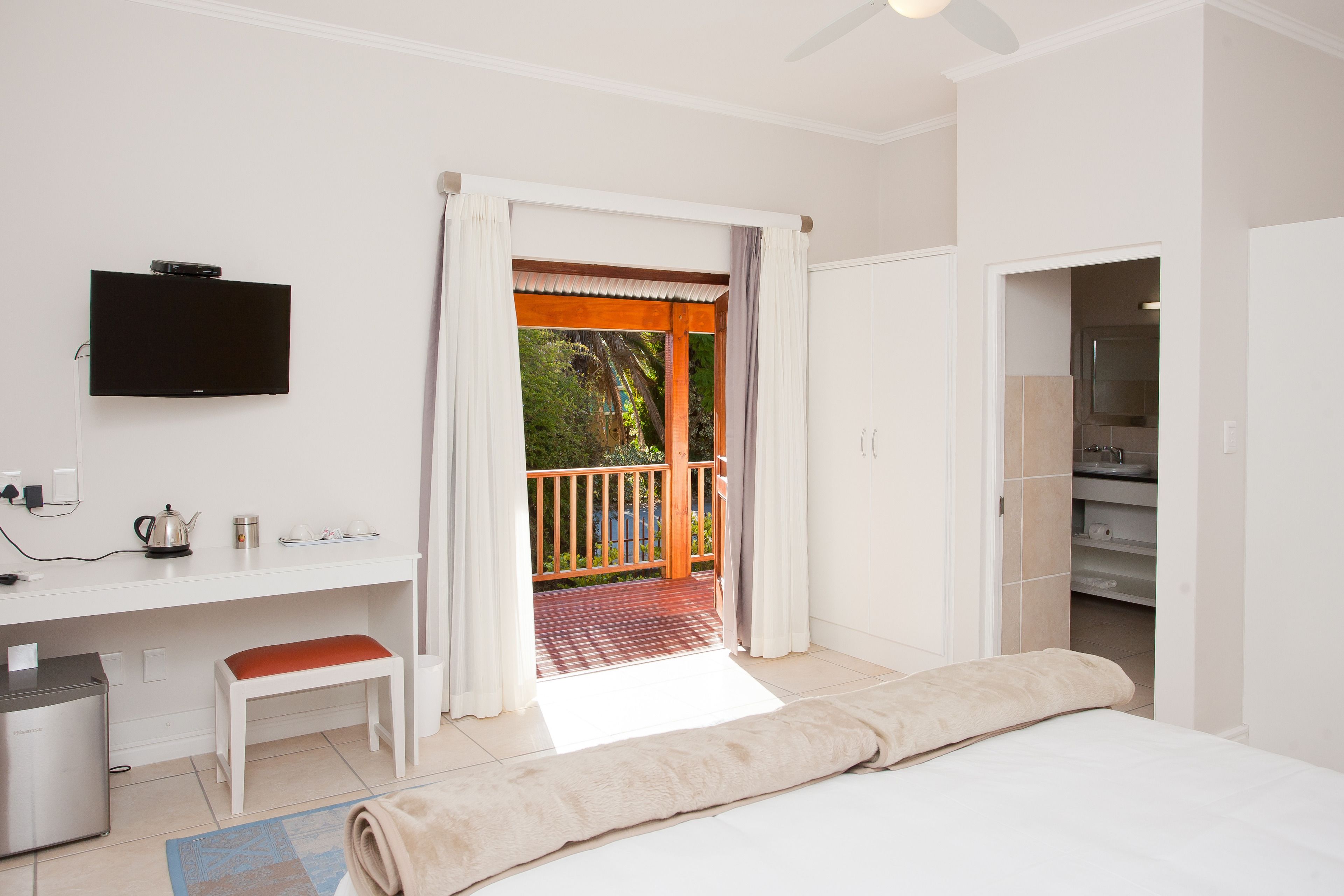 Un dormitorio con cama, escritorio con televisor, baño y balcón con vistas a zonas verdes.