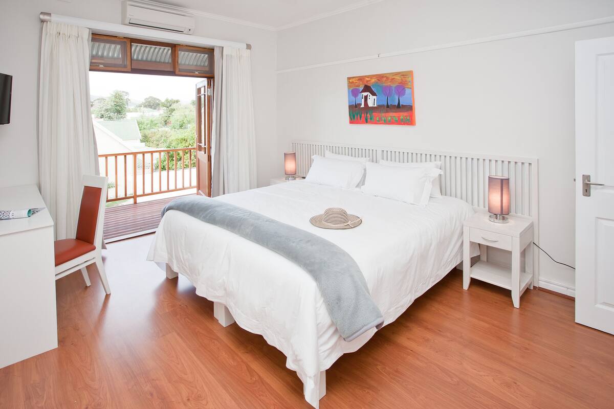 A bedroom with a large bed, white curtains, a painting, a bedside table with a lamp, and a view of greenery outside.