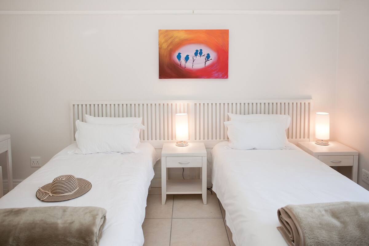 A double room with two beds, a bedside table with a lamp, a wall-mounted artwork, and a striped hat on the bed.