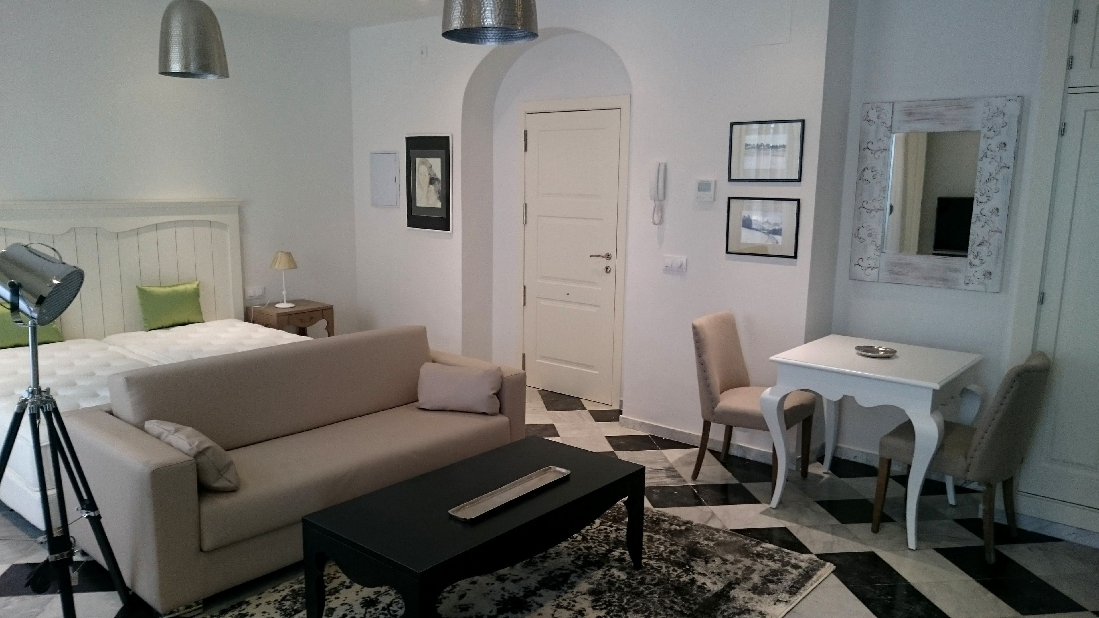 Habitación de hotel con una cama, un sofá, una mesita, un escritorio y una silla. Hay una lámpara, un espejo y cuadros en las paredes.