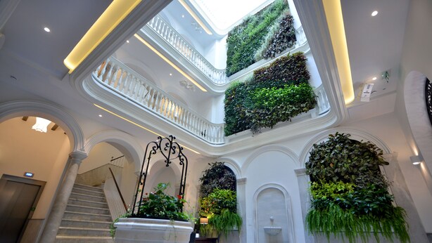 Un atriumo de varios niveles con una escalera central, columnas blancas y jardines verticales verdes.