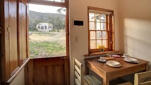 A room with a wooden table set for dining, a window with a view of a building and trees, and a door leading outside. - Keurfontein Country House (Willowmore)