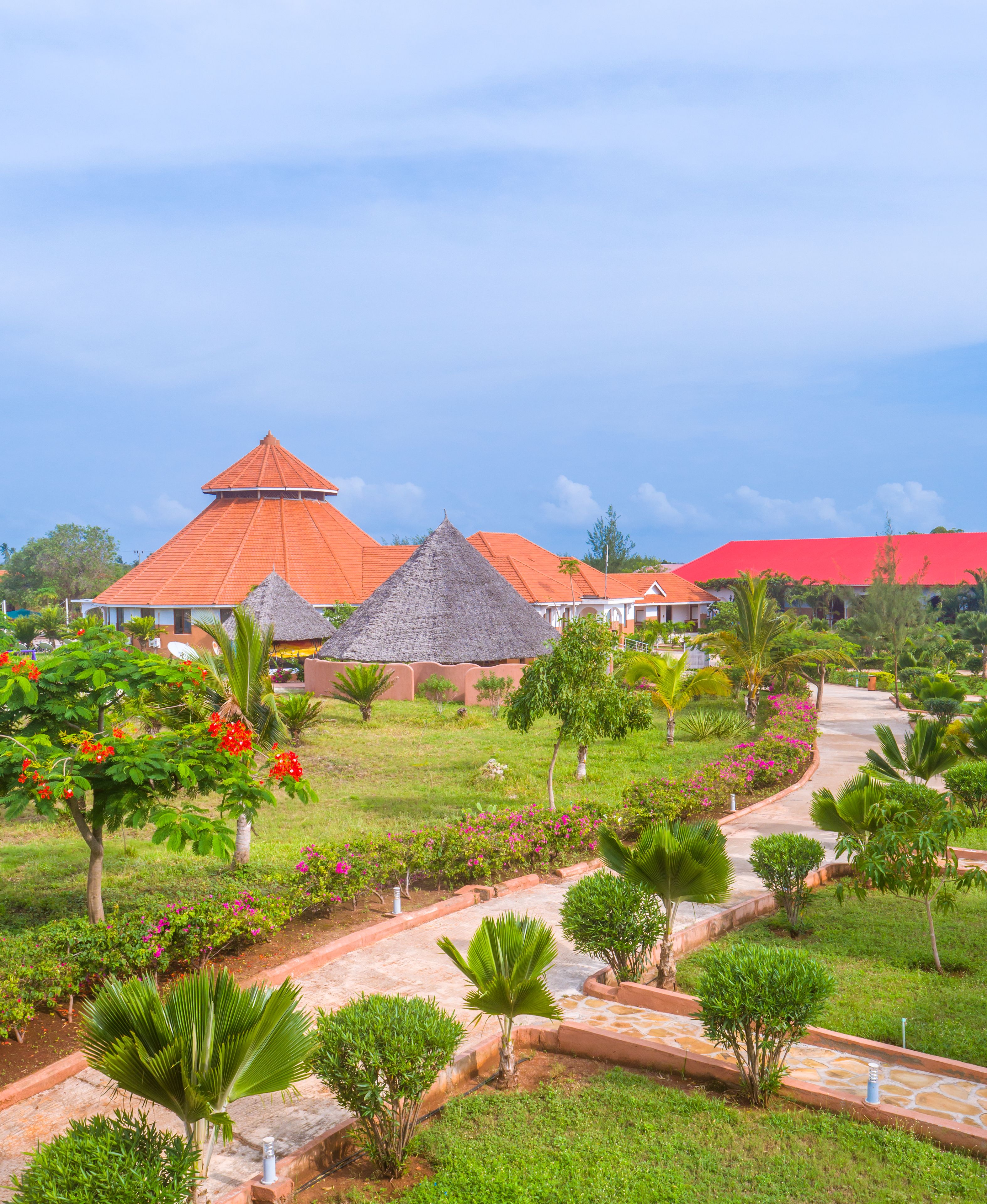 Um resort com edifícios de telhado de palha, um jardim bem cuidado com diversas plantas e flores, e um calçadão pavimentado.