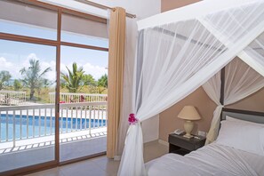 Habitación de hotel con ventana grande, cama, mesita de noche y vista a una piscina y palmeras.