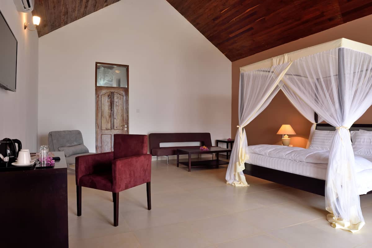 A bedroom with a four-poster bed, a red chair, a grey armchair, a wooden ceiling, and a wall-mounted TV.