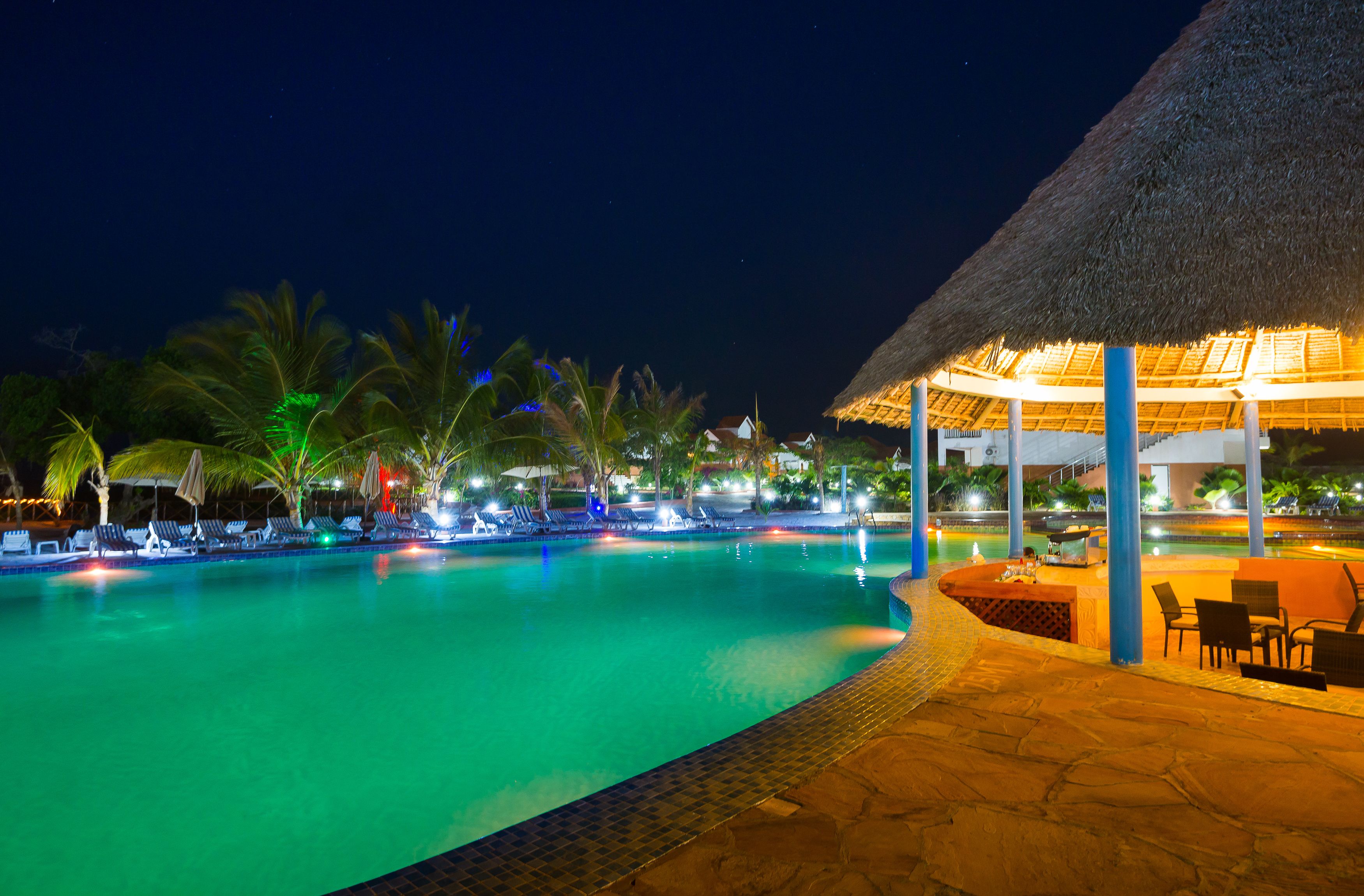 A nighttime swimming pool with lounge chairs and a thatched-roof structure.