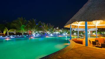 A nighttime swimming pool with lounge chairs and a thatched-roof structure.