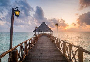 Un muelle de madera que lleva a una estructura de techo de paja con faroles, con vista a un mar tranquilo al atardecer.