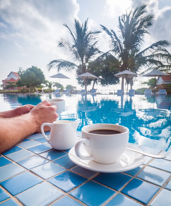 A poolside table with a cup of coffee and a cup of tea, palm trees, and a house in the background.