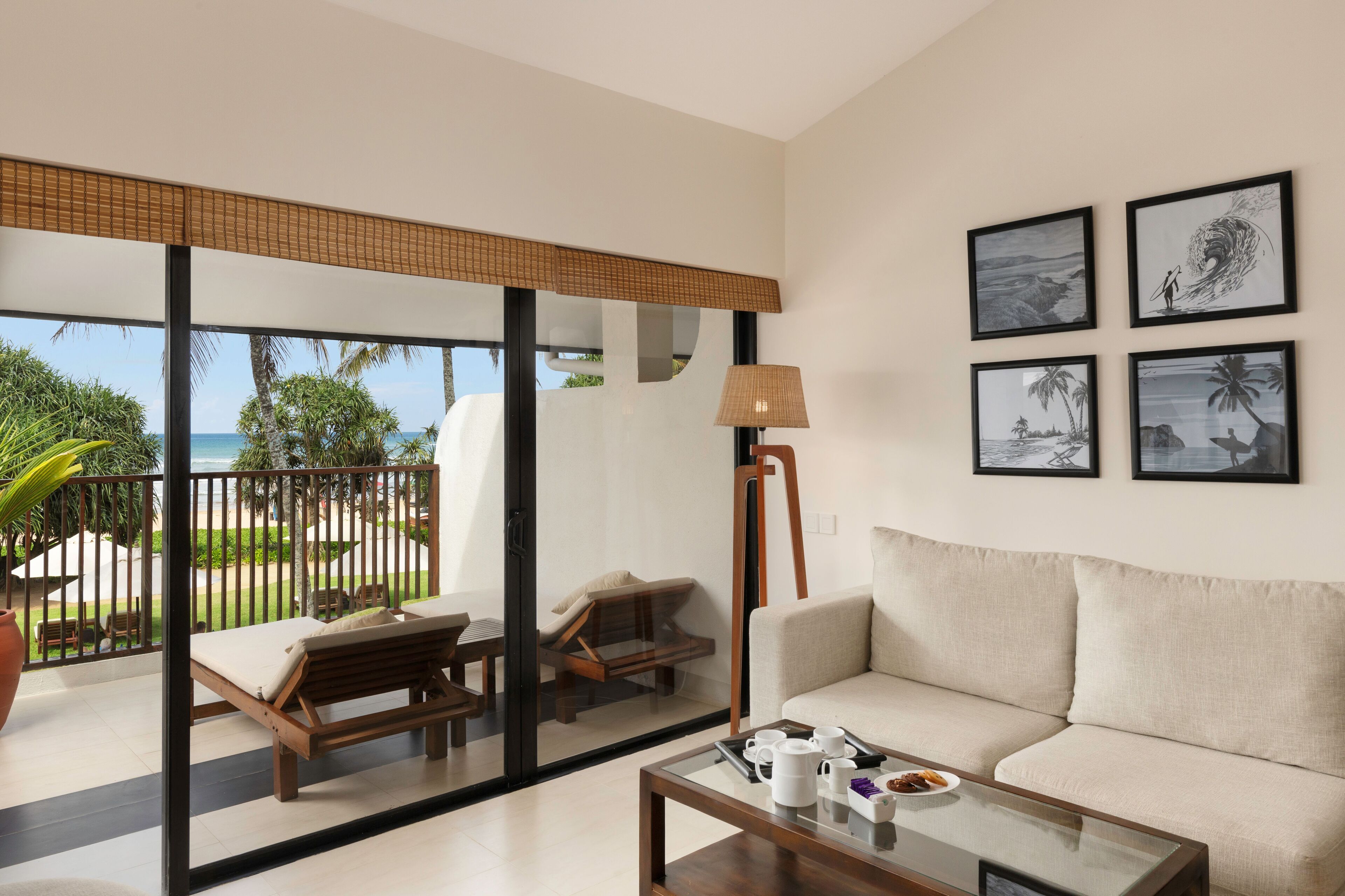 A modern living room with a sofa, a coffee table, and a view of the ocean through large glass doors. There are framed pictures on the wall and a lamp.