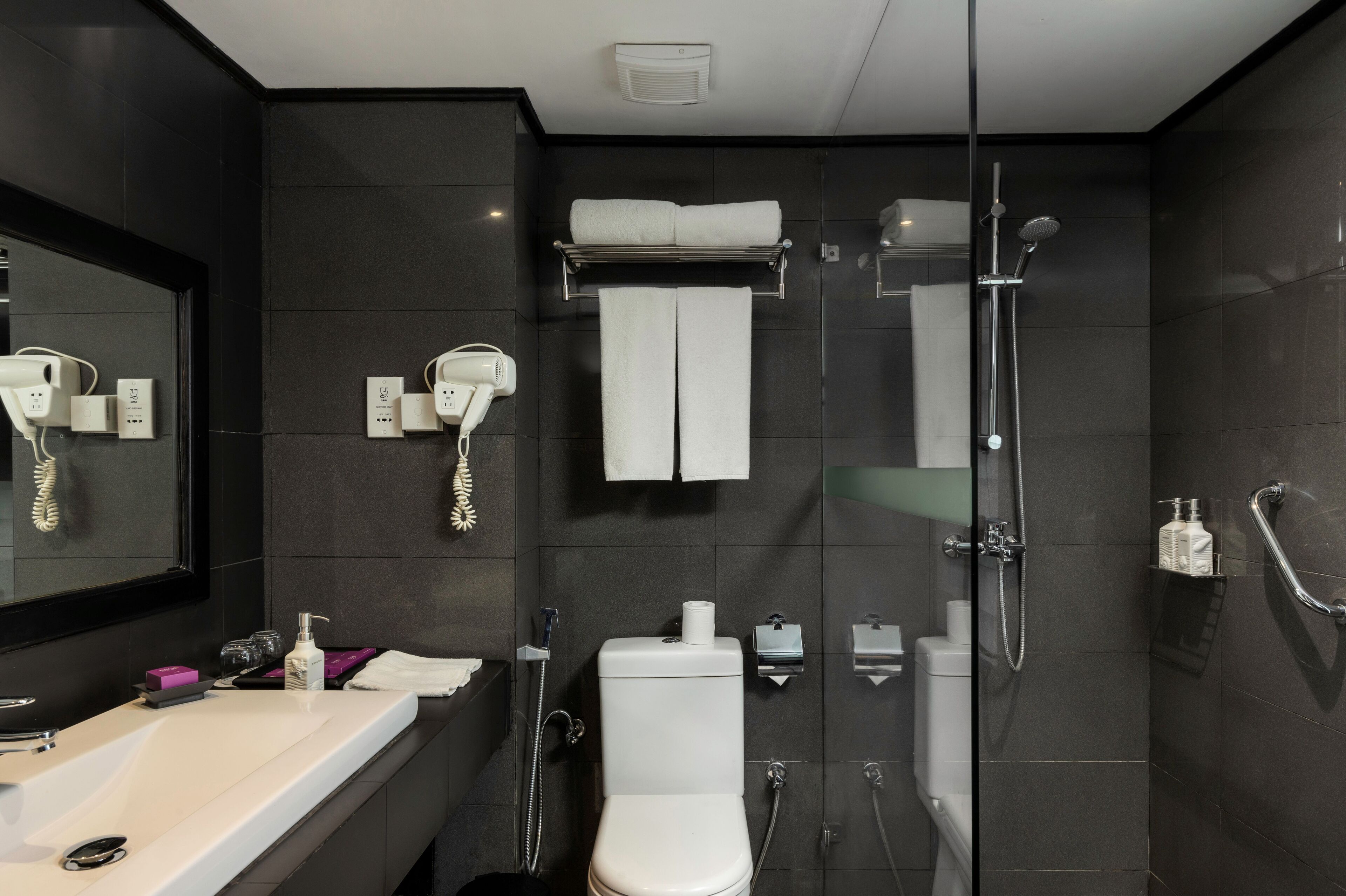 Un baño moderno con un lavabo blanco, inodoro, ducha con puerta de cristal y espejo con un atril para teléfono y toallero.