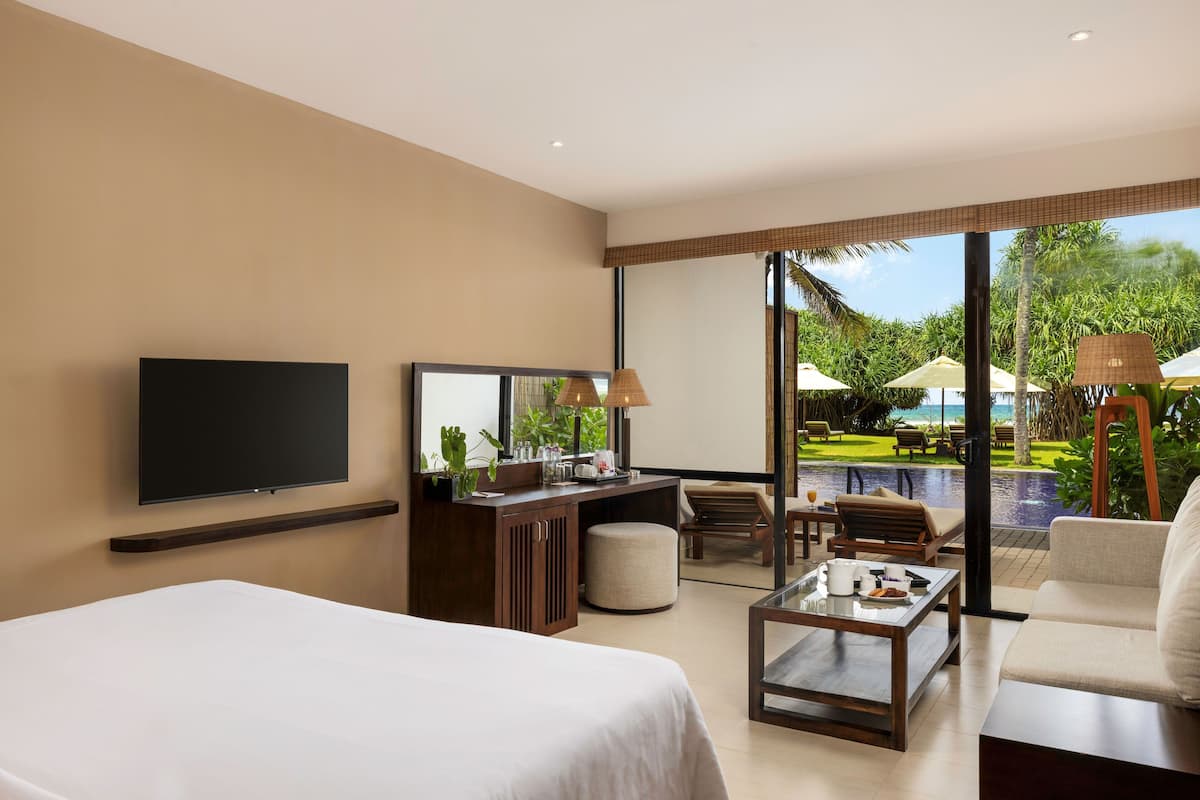 A hotel room with a bed, a desk with a TV, a coffee table, and a seating area with a view of a pool and greenery.