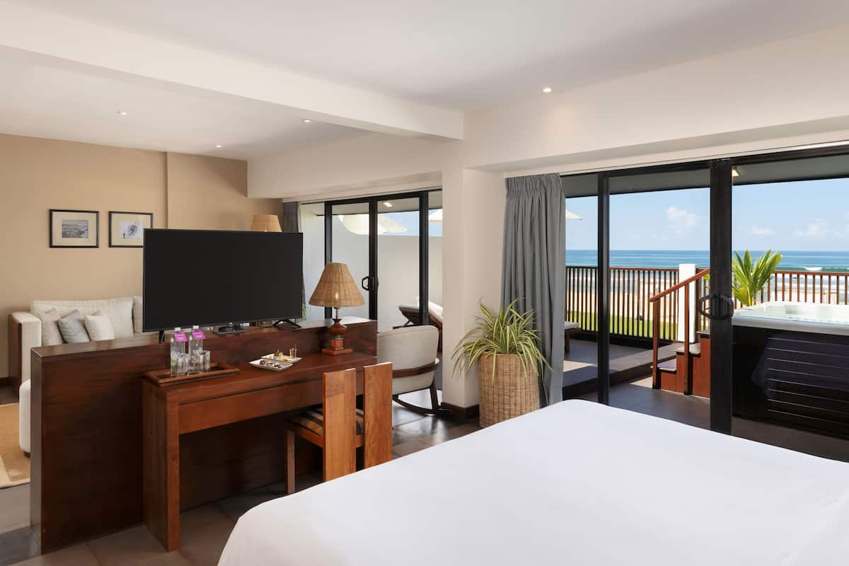 A hotel room with a large bed, a desk with a TV, a seating area, and a balcony with a view of the ocean.