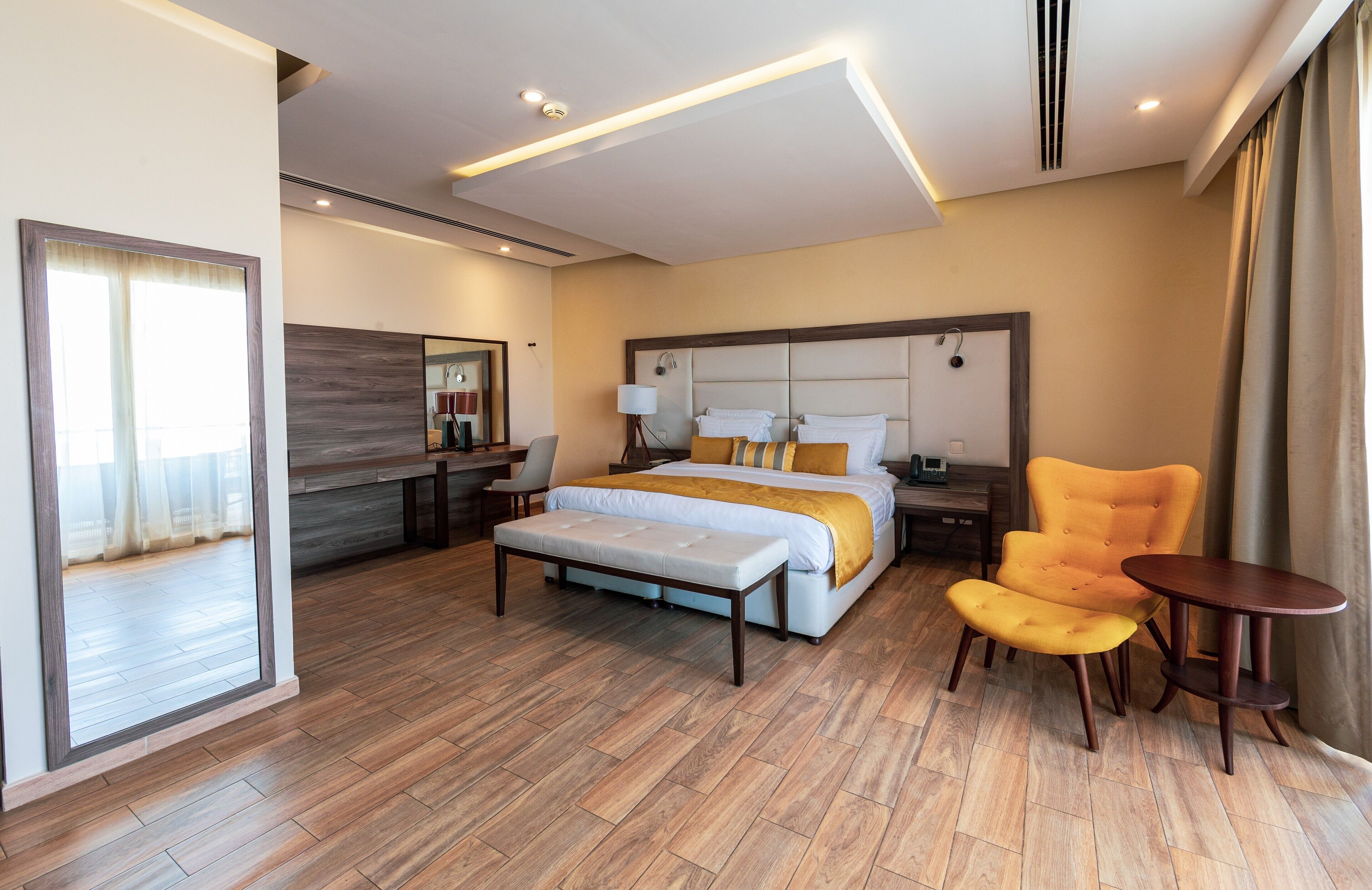 Quarto de hotel moderno com uma cama grande, mesa de cabeceira, uma cadeira, uma mesinha e um espelho.