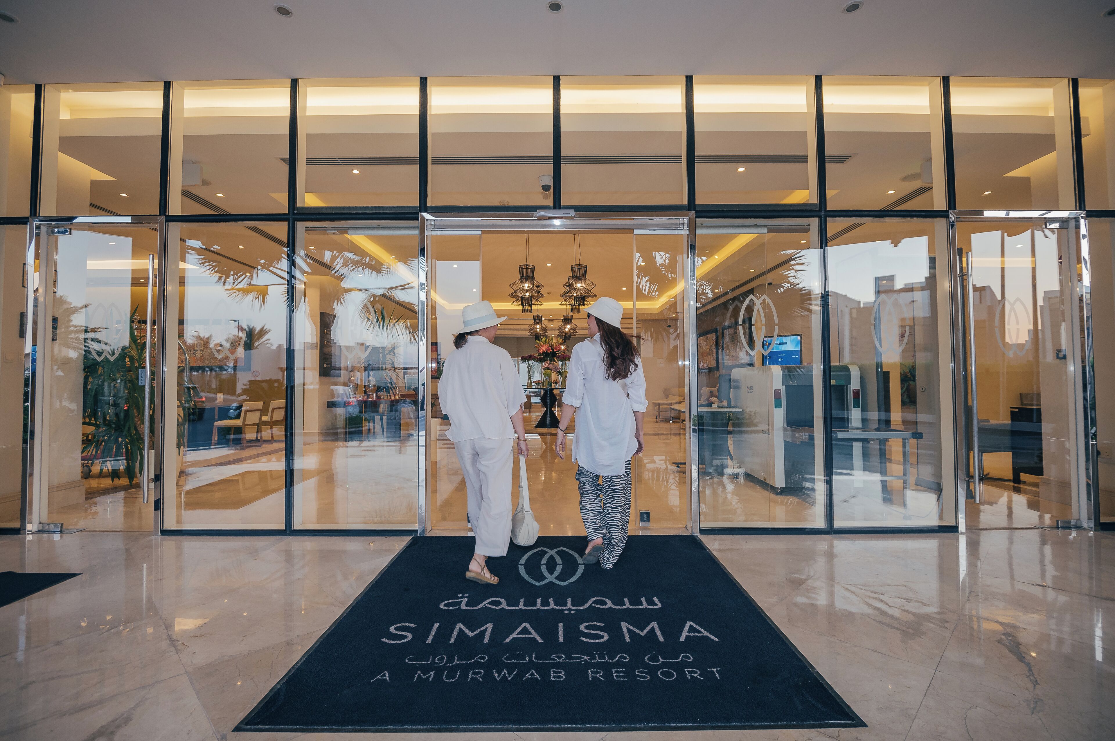A hotel entrance with a glass facade, a black mat with the hotel's name, and a chandelier visible inside.
