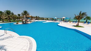 Une grande piscine d’un bleu éclatant, aux bords ondulés, entourée de chaises longues et de parasols, avec des palmiers et un bâtiment en arrière-plan.
