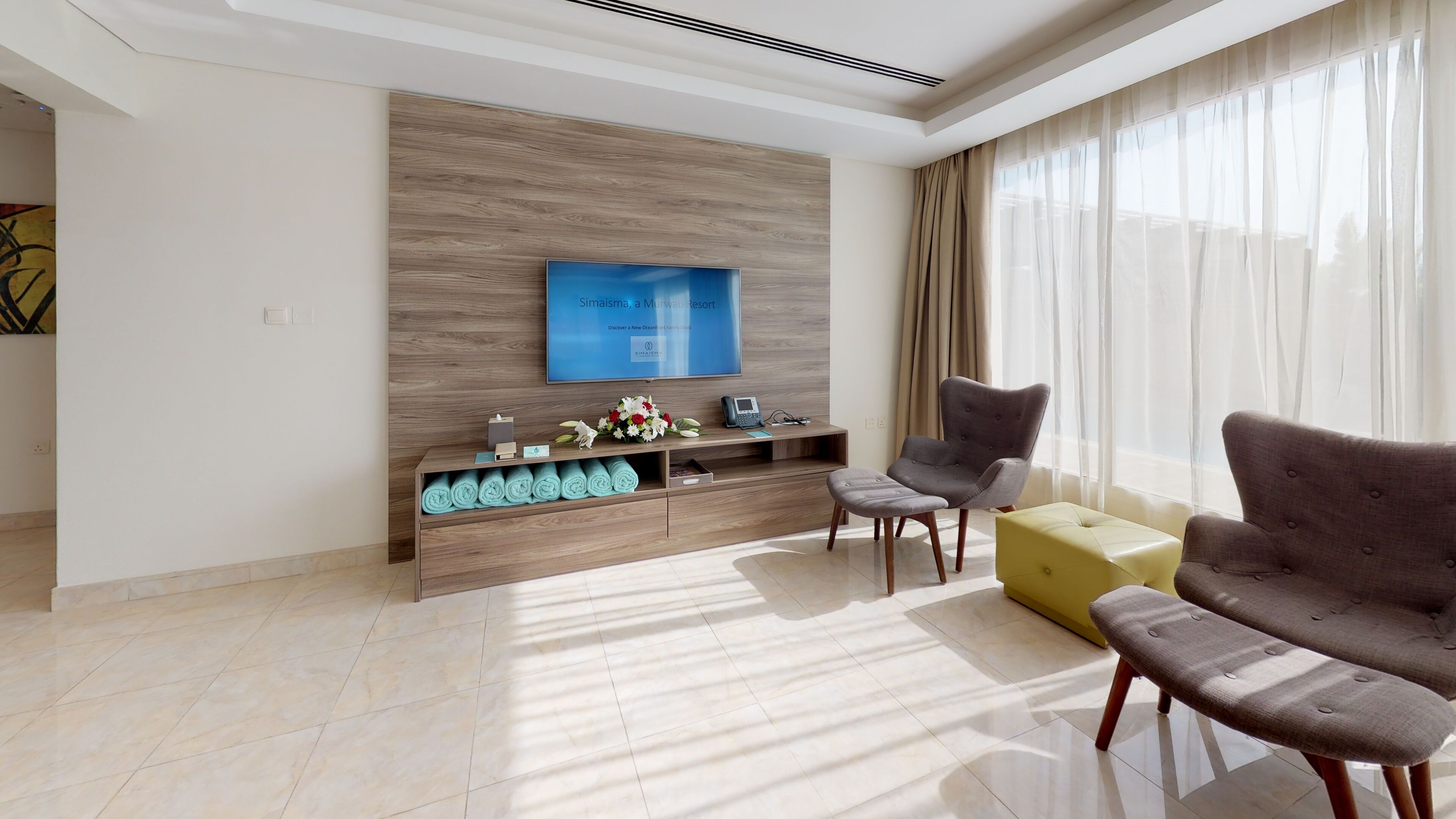 A modern living room with a flat-screen TV mounted on a wooden wall, a sofa, a chair, and a small table with a phone and decorative items.