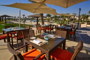 Des tables à manger en plein air, accompagnées de chaises en osier et de coussins orange, disposées sous de grands parasols, avec une piscine et des palmiers en arrière-plan.