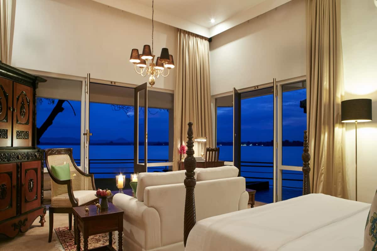 Une chambre d’hôtel avec un grand lit, deux fauteuils, une armoire en bois et une vue sur l’eau au crépuscule.