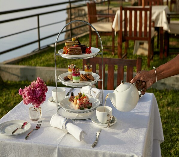 A table set for a tea service with a three-tiered dessert stand, a vase of pink flowers, and a view of a body of water in the background.