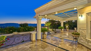 A covered outdoor area with a view of the city at dusk, featuring a tiled floor, potted plants, and a pergola structure. - Frido Luxury Villa (Zakynthos)