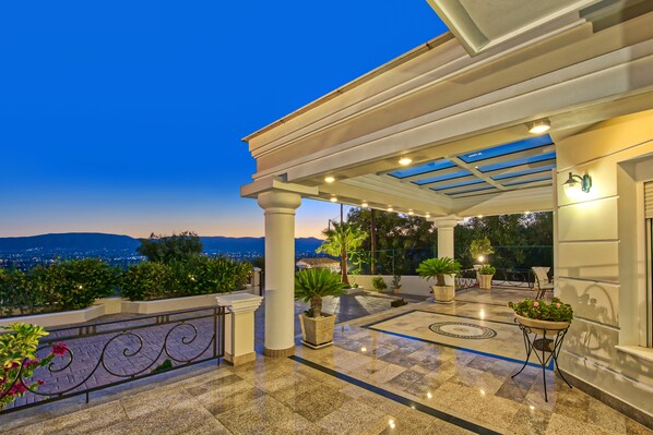 A covered outdoor area with a view of the city at dusk, featuring a tiled floor, potted plants, and a pergola.