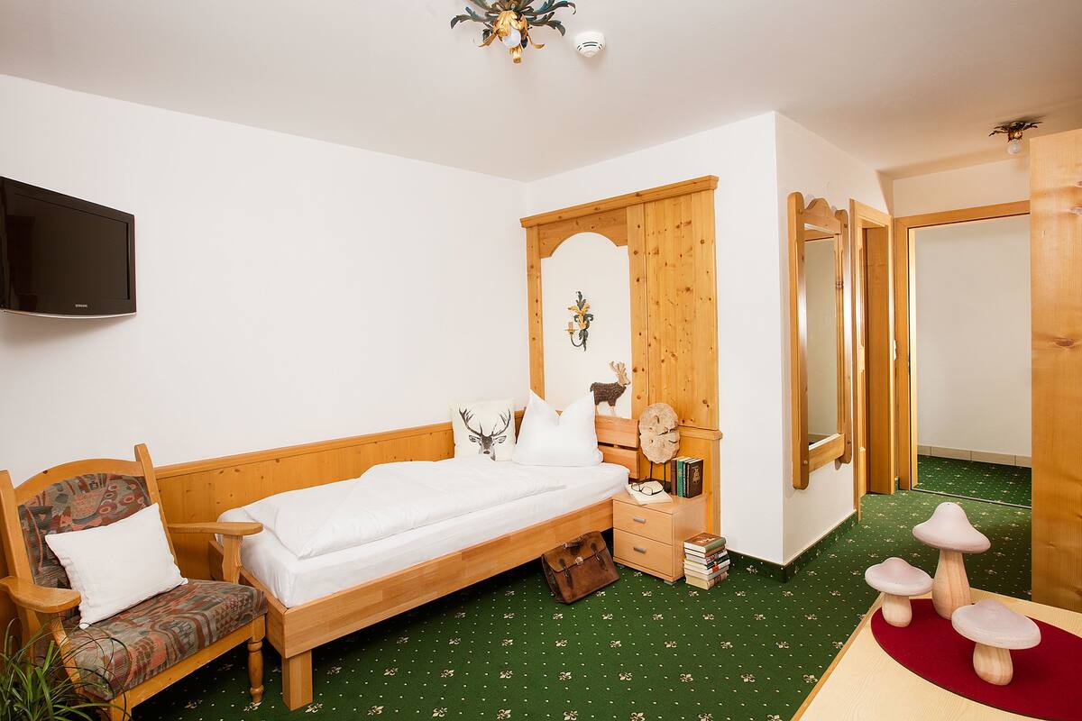 A room with a bed, a chair, a wooden wardrobe, a mirror, and a television on the wall.