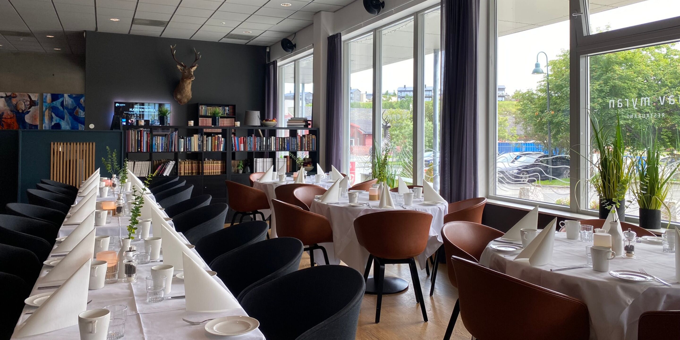 Un restaurante con una mesa larga dispuesta para comer, una estantería y un amplio ventanal con vistas al exterior.