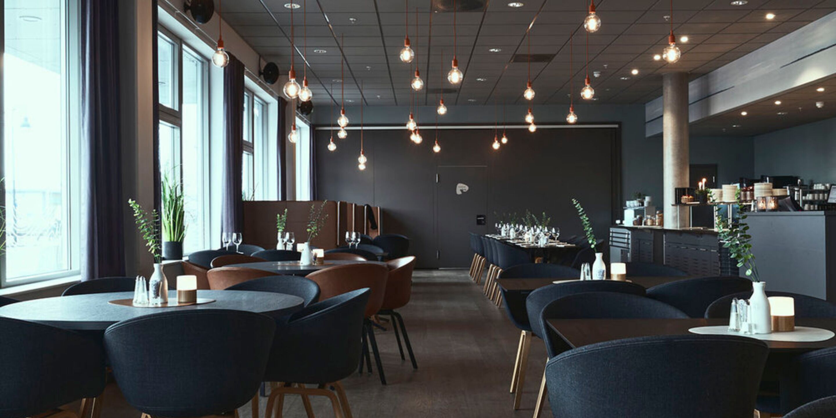 A modern restaurant interior with round tables, dark chairs, hanging pendant lights, and a bar area.