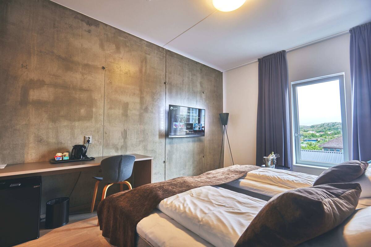 Ein modernes Hotelzimmer mit einem großen Bett, einem Schreibtisch mit Stuhl, einem Fernseher und Blick ins Freie.