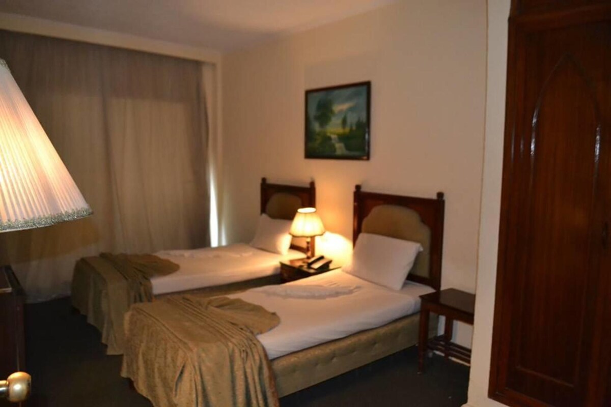 A hotel room with two beds, a nightstand with a lamp, a painting on the wall, and a wooden door.