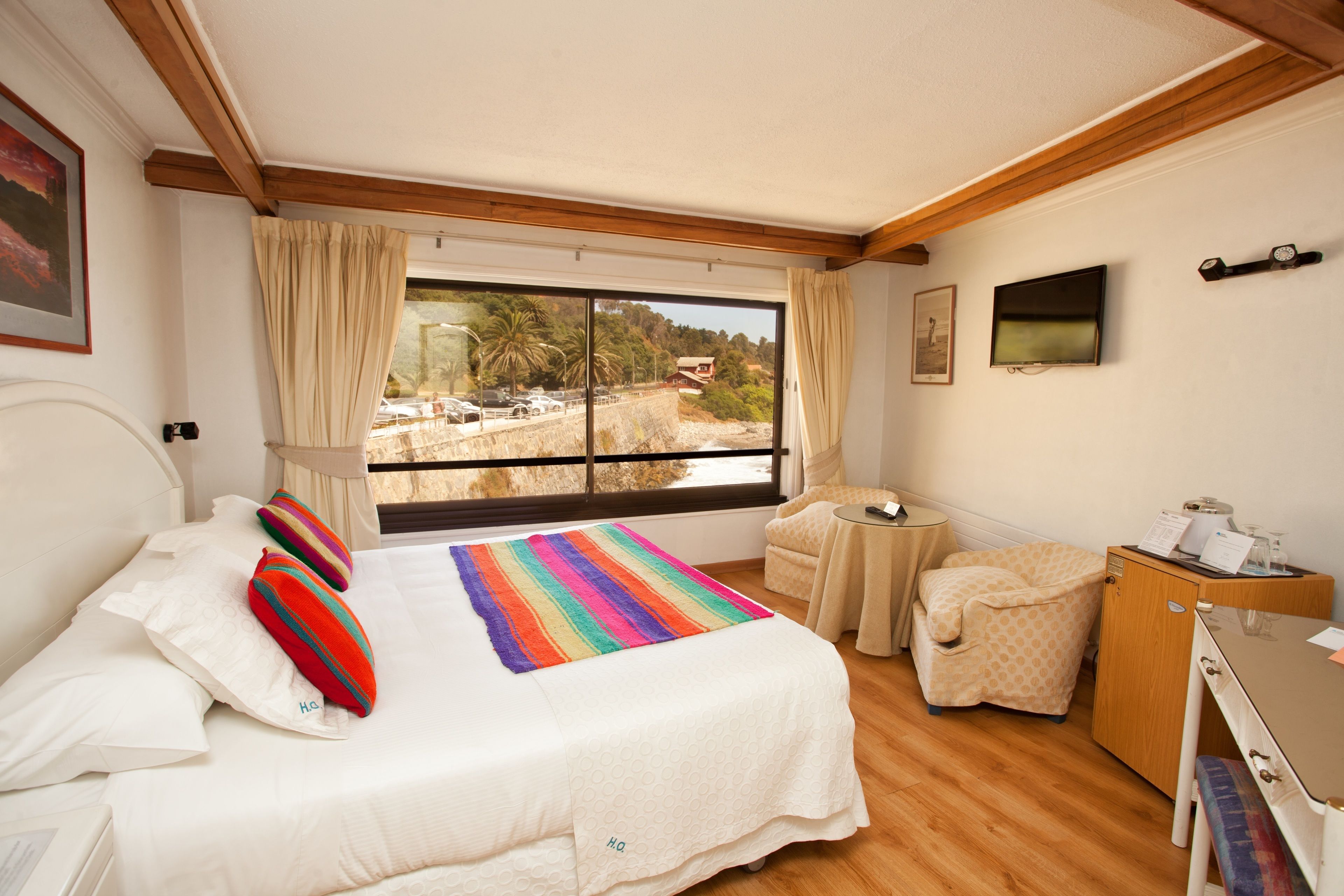 A bedroom with a large bed, a desk, a chair, a TV, and a window with a view of a beach and buildings.