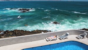 Piscina com água azul cristalina, rodeada por espreguiçadeiras, com vista para um litoral rochoso onde as ondas quebram.
