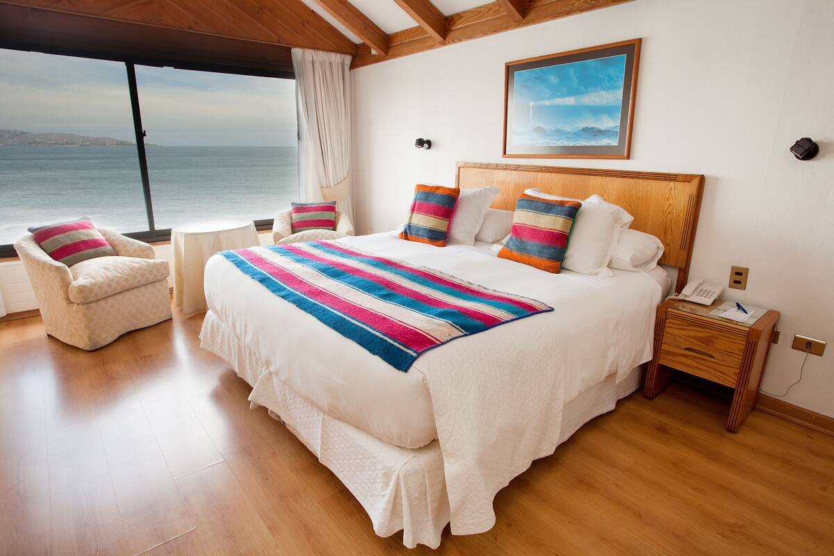 Un dormitorio con una cama grande, una mesita de noche de madera, una silla y vista al mar.